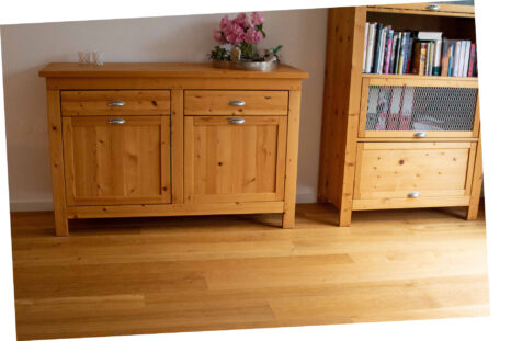 Heller Echtholzparkettboden vor weißer Wand und farblich passender heller Holzkommode und Holzschrank. Auf der Kommode steht eine Blumenvase, im Schrank sind diverse Ordner.