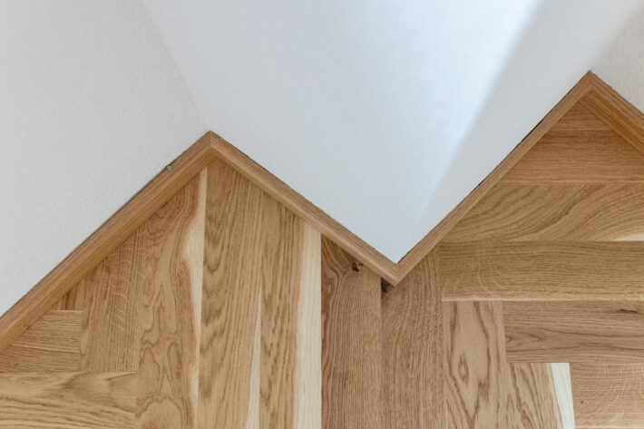 Blick von oben auf Fischgrätparkett an Wand, drei Ecken, Sockelleiste in Parkettoptik, weiße Wand.