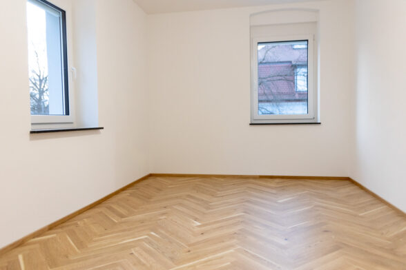 Blick in viereckigen Raum komplett in Fischgrätparkett, weiße Wände, zwei Fenster im Bild.