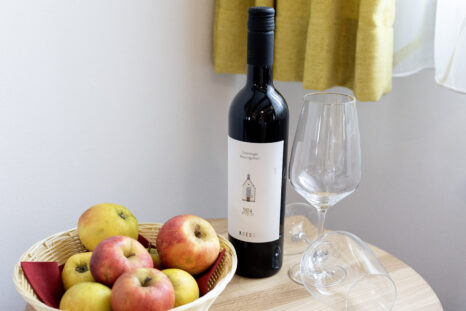 Blick auf Beitisch in Stilllebenoptik, Schale mit Äpfeln, Weinflasche, Weinglas, vor heller weißer Wand und gelben Vorhängen.