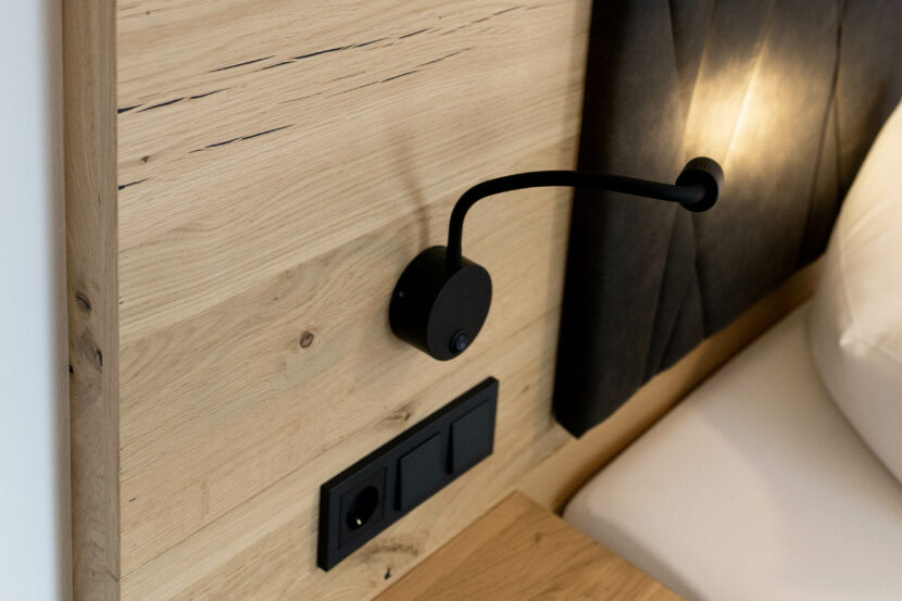Blick auf holzvertäfelte Rückwand neben Bett, schwarzer Steckdosenleiste, schwarze Nachttischlampe.