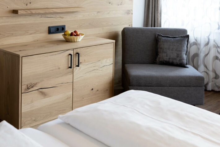 Eckansicht Zimmer Hofgut Schellenberg, im Vordergrund weiße Bettwäsche, links im Bild heller Naturholzschrank, rechts daneben schwarzgrauer Sessel ohne Lehnen, daneben helle Vorhänge.