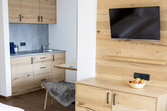 Hotelzimmer Hofgut Schellenberg, rechts im Bild Naturholzkommode aus hellem Holz, holzgetäfelte Wand mit Flachbildfernseher, links im Bild Einblick in die Küchenzeile, Küchenschränke sind in der gleichen hellen Naturholzoptik gehalten. Die Wände sind weiß verputzt, der Boden aus hellem Vinyl.