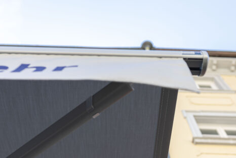 Blick von direkt unten auf weiße Markise mit anthrazit-grauer Unterseite, weißer Volant mit blauer Firmenbeschriftung.