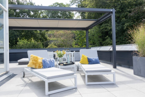 Q-bus mit dunklem Metallrahmen über weiß gefliester Terrasse, hellbeiges Segel halb eingefahren. Zwei weiße Liegen mit blauem und gelbem Kissen, Bäume im Hintergrund.