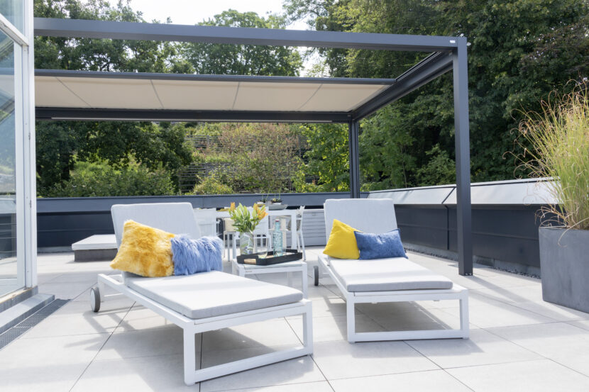 Q-bus mit dunklem Metallrahmen über weiß gefliester Terrasse, hellbeiges Segel halb eingefahren. Zwei weiße Liegen mit blauem und gelbem Kissen, Bäume im Hintergrund.