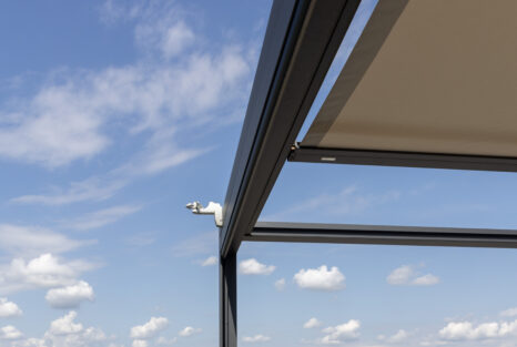 Blick von unten auf linke Seite von Q-Bus, schwarzer Metallrahmen vor blauem Himmel, beiges Segel ausgefahren.