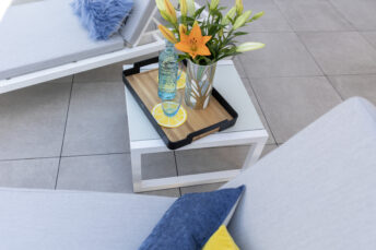 Blick von oben auf weißes Tischchen auf weiß gefliester Terrasse, Tischchen steht zwischen zwei weißen Liegen. Auf dem Tisch steht ein Tablett mit Wasserglas, Wasserflasche und Blumenstrauß in Vase.