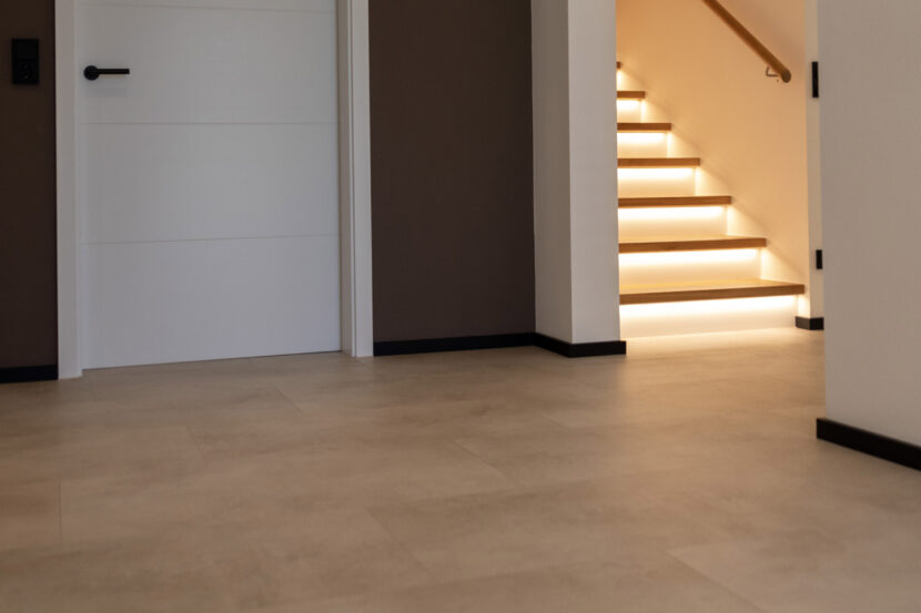 Bild zeigt Flur mit Mineralboden, links im Bild weiße Türe vor brauner Wand, rechts beleuchtete weiße Treppe ins Obergeschoss.