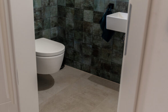 Heller Mineralboden in WC, seegrüne Fliesenwand, weißes Waschbecken und weißes Klosett.