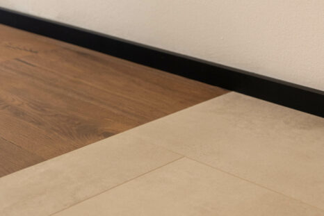 Übergang Mineralboden zu Echtholzparkett, schwarze Sockelleiste und weiße Wand.