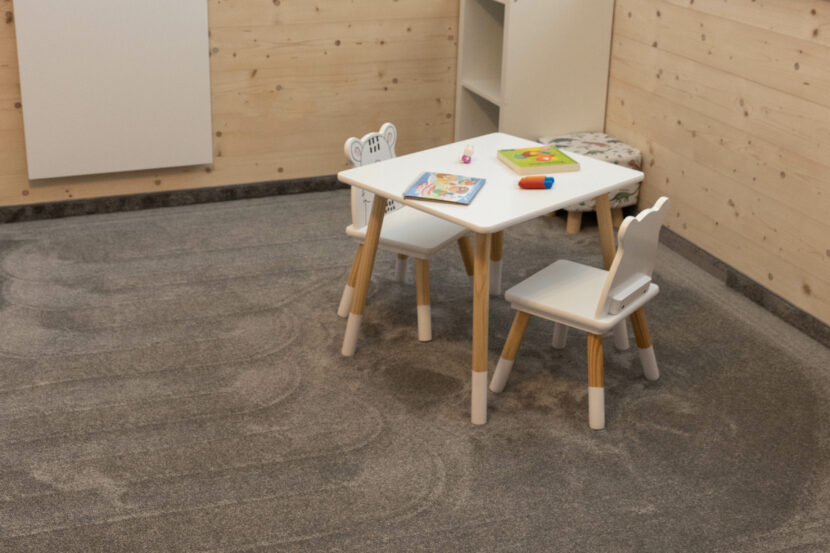 Blick auf Ecke eines Kinderzimmers, der Teppich ist dunkel und flauschig, in der Bildmitte sind ein weiß lackierter Kindertisch aus Naturholz mit zwei kleinen Stühlen zu sehen, die Wand im Hintergrund ist mit hellem Naturholz getäfelt. In der Raumecke steht ein weiß lackiertes, offenes Regal.