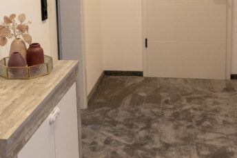 Bild von schräg oben auf neuen, dunklen flauschigen Teppich, links im Vordergrund Naturholzkommode mit weißen Türen, im Hintergrund weiße Wände mit weißer Tür.