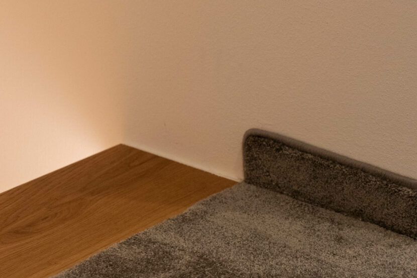 Raumecke, neuer, dunkler Teppich mit Teppichsockel neben Parkettboden vor weißer Eckwand.