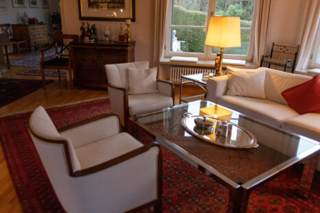 Frisch aufgepolsterte weiße Sessel in Wohnzimmer neben Glastisch auf rotem Teppich, im Hintergrund weißes Ecksofa mit rotem Kissen, Parkettboden.