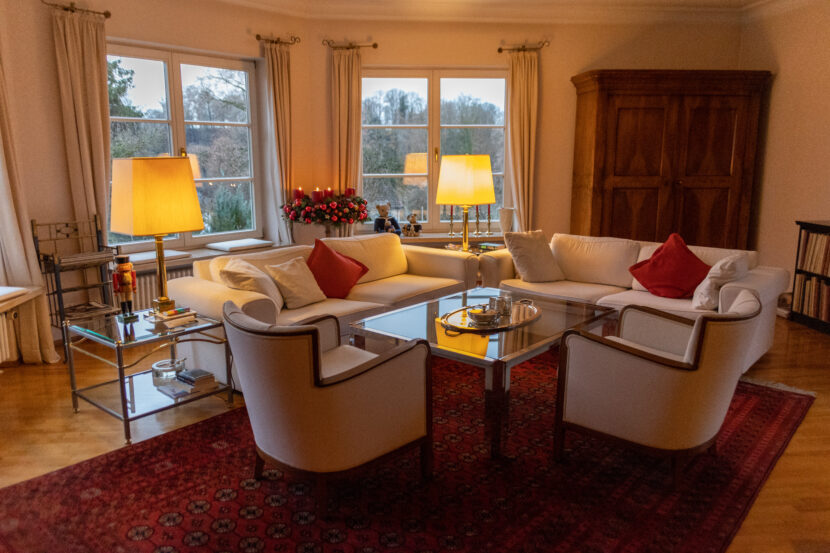 Bild zeigt zwei frisch aufgepolsterte weiße Sessel in Wohnzimmer auf rotem Teppich um Glastisch, zwei weiße Ecksofas im Hintergrund, weiße Wände und zwei angeschaltete Stehtischlampen auf Glastischen