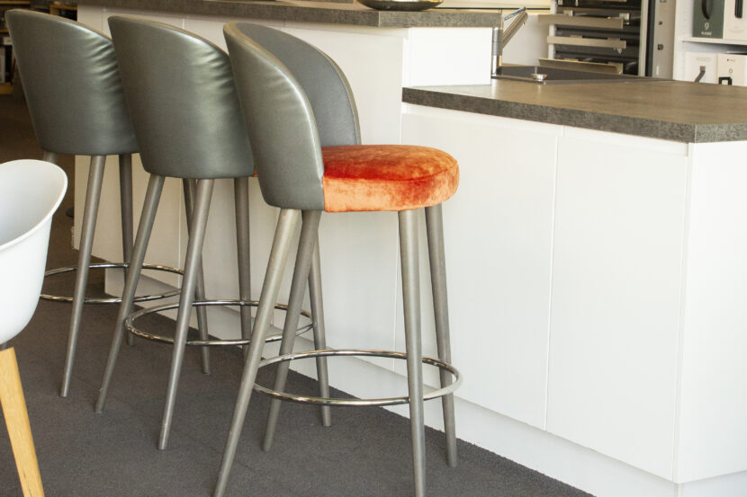 Bild von der Seite von drei frisch gepolsterten Barhockern, Beine aus hellem Metall, dunkelgraue Lehnen, beim Hocker rechts vorne ist das orangene Velourspolster zu sehen.