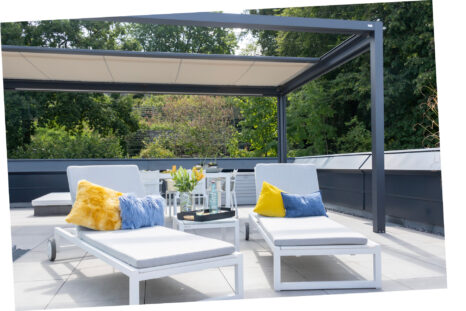 Q-bus mit dunklem Metallrahmen über weiß gefliester Terrasse, hellbeiges Segel halb eingefahren. Zwei weiße Liegen mit blauem und gelbem Kissen, Bäume im Hintergrund.