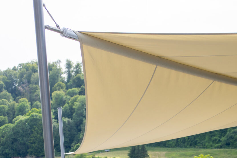Helles beiges Sonnensegel, aufgespannt an hellem Metallrahmen, Wald im Hintergrund.