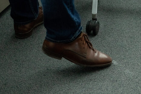 Nahansicht neue Teppichfliesen in hellem blaugrau, mittig im Bild Füße in braunen Lederschuhen.