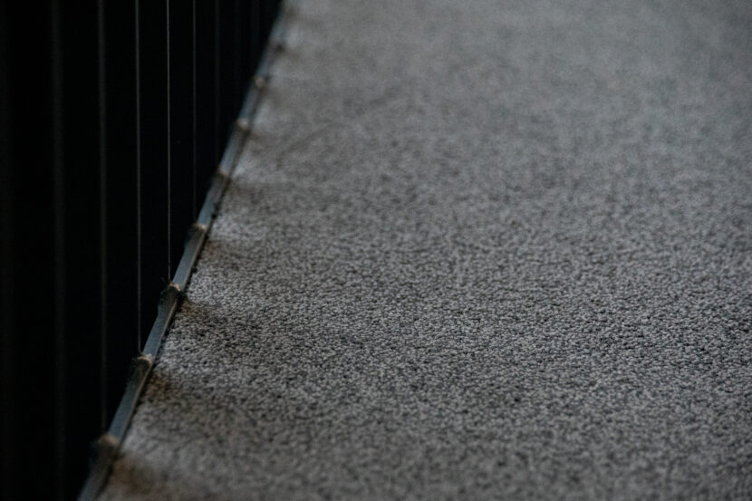 Graue Teppichfliesen in Nahaufnahme, links schwarzes Metallgeländer.