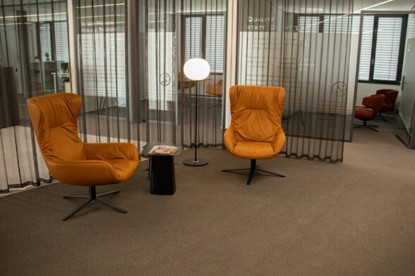 Büroflur mit hellbraunen Teppichfliesen, im Hintergrund mehrere Büros, im Vordergrund zwei sonnengelbe Polstersessel mit Stehlampe.