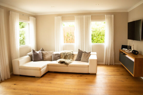 Frisch eingerichtetes Wohnzimmer, Fußboden aus österreichischem Echtholzparkett, in der Raummitte ein weißes Ecksofa, weiße Gardinen vor weißen Wänden.