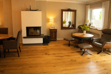 Frisch renoviertes Esszimmer, österreichisches Echtholzparkett, rechts im Bild weiße Gardinen, mittig weißer Kamin und Spiegel. Mehrere kleine Holztische mit Stühlen im Hintergrund.