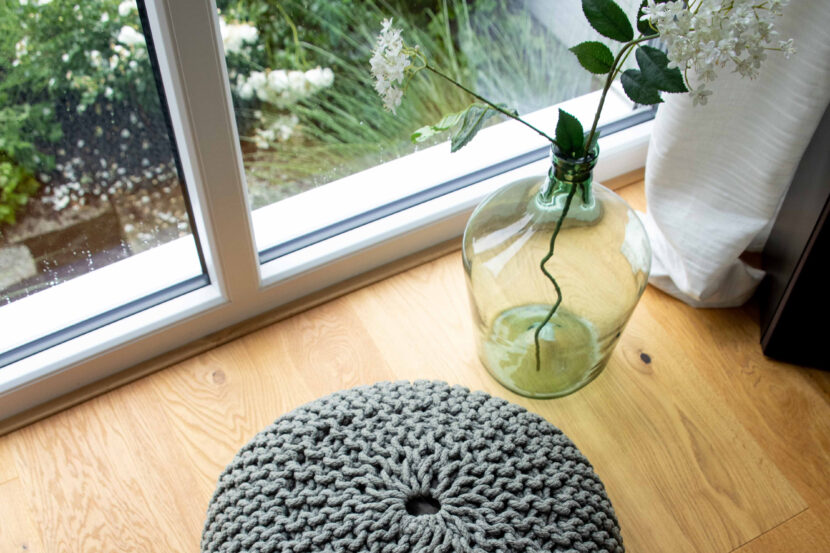 Echtholzparkett von oben. links weiße Fensterfront mit Blick auf Garten, im Vordergrund Strickkissen.