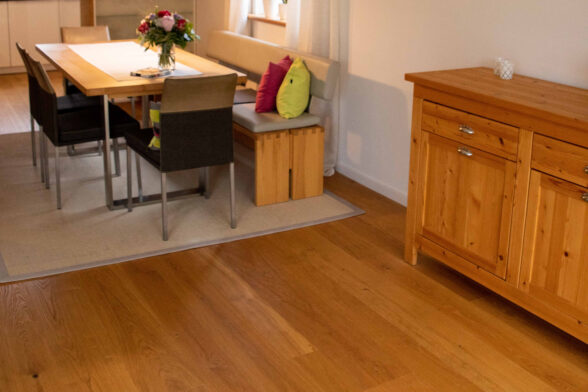 Schrägansicht Esszimmer, neues Echtholzparkett, rechts im Bild Holzkommode im passenden Ton, links im Bild weißer Teppich mit Esstisch und drei Stühlen mit Sitzbank. Weiße Wände.