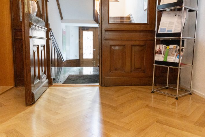 Frisch verlegter Fischgrätparkett aus hellem Holz, Ansicht aus Bodenperspektive Richtung Ausgang zur Treppe. Rechts im Bild steht ein Metallregal vor weißer Wand.