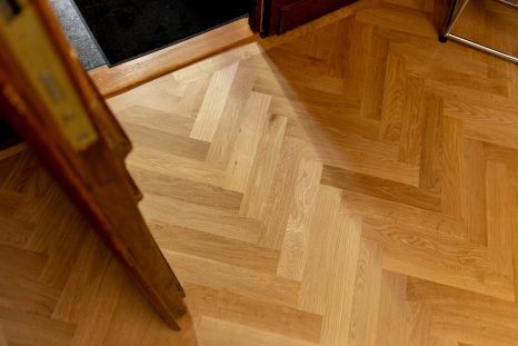 Neu verlegter Fischgrätparkett aus hellem Holz aus der Vogelperspektive und im Detail. Aus der offenen Tür fällt ein Streifen Licht auf das Parkett.
