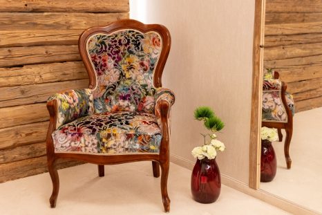 Totalaufnahme frisch gepolsterter Lehnenstuhl aus Holz mit floralem Samt vor Spiegel, Holzwand und Blumenvase.