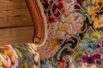 Nahaufnahme des frisch gepolsterten Armlehnenstuhls mit floralem Stoff. Sichtbar sind eine Armlehne, ein Teil der Rückenlehne, vor dem Hintergrund einer Holzwand