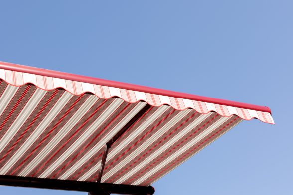 Ansicht von unten und vorne einer rot-weiß gestreiften Kassettenmarkise, ausgefahren mit rot lackierter Front vor blauem Himmel.