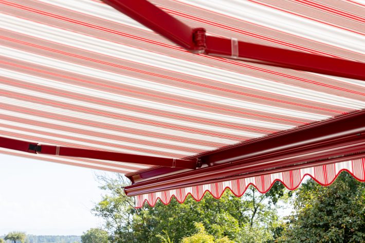 Seitenansicht einer ausgefahrenen Kassettenmarkise mit rot weiß-gestreiftem Stoff, Gelenkarme und Front sind rot lackiert, der Volant ist eingezogen. Im Hintergrund sind blauer Himmel und grüne Bäume zu sehen.