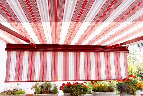 Sicht von Balkon aus auf ausgefahrene, rot-weiß gestreifte Kassettenmarkise, am unteren Bildrand sind verschiedene Blumentöpfe sichtbar. Der Volant ist heruntergelassen.