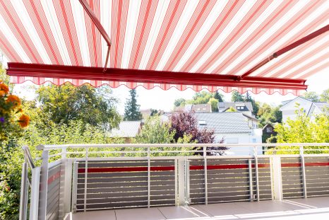 Ansicht von innen auf ausgefahrene Kassettenmarkise und Balkon, die Markise ist rot-weiß gestreift, die Gelenkarme und die Front sind rot lackiert. Der Balkon hat ein weißes Geländer, graue Fließen und eine hellgraue Holzverkleidung. Der Volant ist eingefahren.