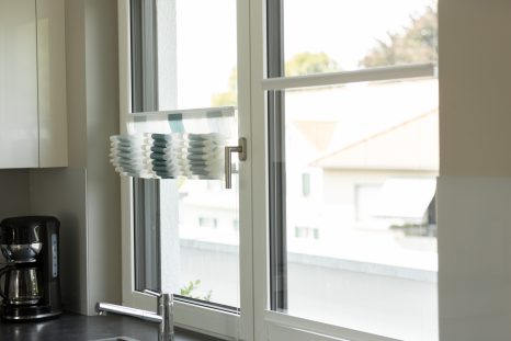 Schräge 3/4 Ansicht von Doppelfenster in der Küche mit gerafften, blau-weiß gestreiften Aeros. Der Fensterrahmen ist weiß, links auf dem Bild ist eine Filterkaffeemaschine zu sehen.