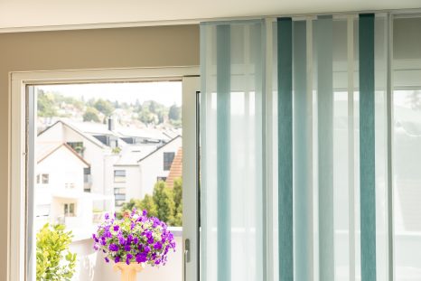 Blau-weiß gestreifte Aeros vor weißem Fenster, das Fenster ist halb verdeckt. Durch das Fenster ist ein Balkon mit Blumenkübeln sichtbar.