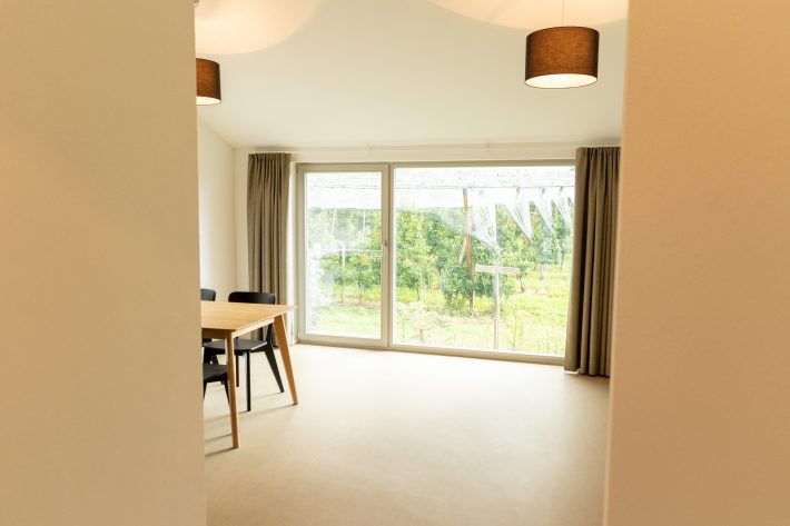 Sicht auf Raum mit neuem Linoleumboden und durchgehender Fensterfront mit hellbraunen Vorhängen von Eingang aus.