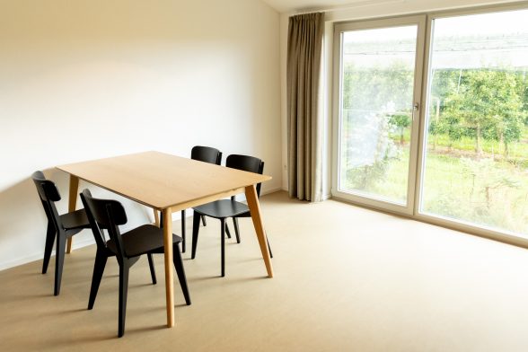 Dreiviertel Ansicht Zimmer mit hellem Linoleumboden, große Fensterfront mit schallschluckendem Vorhang, links im Bild ein heller Holztisch mit vier dunklen Stühlen.