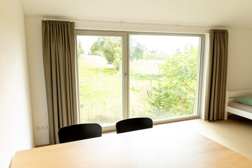 Ansicht Fensterfront frontal mit schalldämpfenden Vorhängen an beiden Seiten, heller Linoleumboden, rechts im Bild ist ein weißes Bett zu sehen, im Vordergrund ein heller Holztisch mit zwei schwarzen Stühlen.