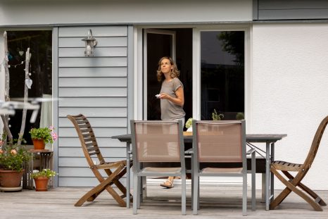 Nahansicht Terrasse unter Q-Bus, zu sehen sind zwei weiße, halb geöffnete Balkontüren und die Hausbesitzerin. Im Vordergrund Gartentisch auf der hellen Terrasse, umgeben von 4 Gartenstühlen.