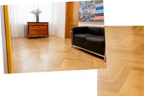 Totale des Zimmers mit frisch verlegtem, hellem Fischgrätparkett. Rechts im Zimmer steht ein schwarzes Sofa, mittig an der Rückwand eine Kommode aus dunklem Holz vor weißem Hintergrund.