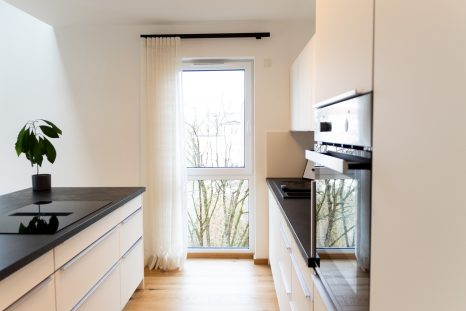 Mitte der Küchenzeile mit bodenhohem Fenster, schwarze Vorhangstange mit creme-farbenem Wellenvorhang, Schiffsdielenboden von Haller Raumgestaltung.