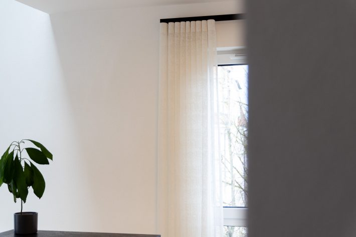 Halb verdecktes weißes Fenster mit schwarzer Deckenschiene und cremeweißen Wellenvorhängen von Haller, Zimmerpflanze auf Tisch links im Bild.