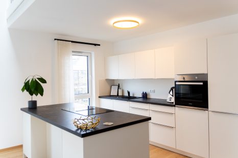 Küchenzeile in weiß mit schwarzem Top, Schiffsdielenboden vor weißer Wand, links im Bild bodenhohes Fenster mit schwarzer Deckenschiene und creme-weißen Wellengardinen.