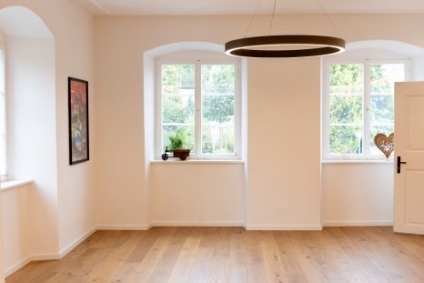 Dreiviertelansicht Pfarrhaussaal, Eichenparkettboden. Weiße Wände, eingelassene Fenster mit Blick auf Bäume. Weißes LED-Ringlicht an der Decke.